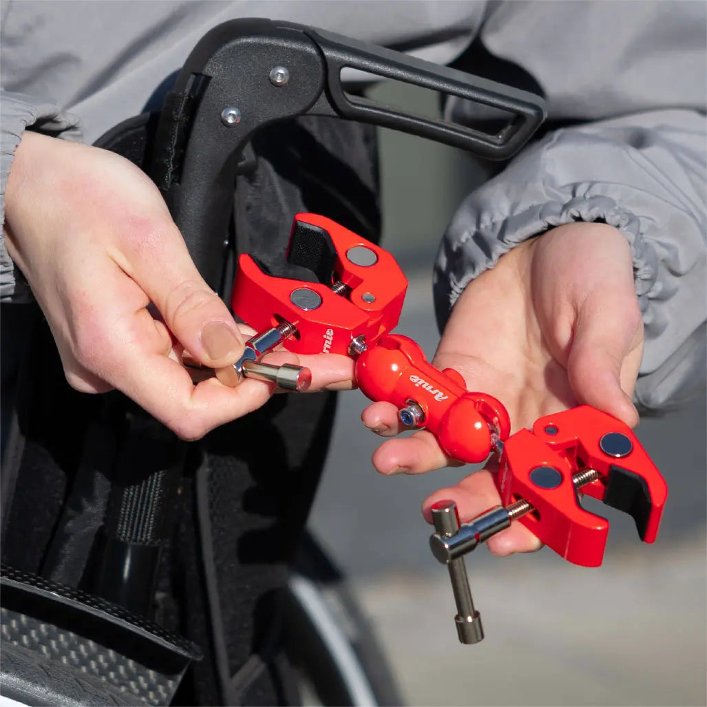 Rote Arnie Anhängerkupplung in der Hand, bereit zur Montage am Rollstuhl. Zubehör für Mobilität und Gepäcktransport.