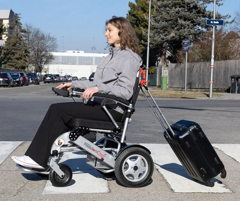 Frau im Rollstuhl mit montierter Arnie Anhängerkupplung, die einen Koffer zieht. Praktisches Zubehör für unterwegs.