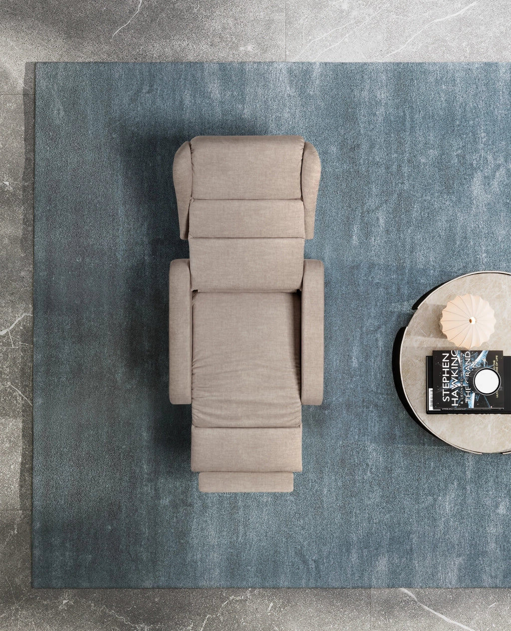 Ein moderner, beige gepolsterter Sessel mit verstellbarer Rückenlehne und Fußstütze, von oben betrachtet. Der Sessel steht auf einem großen, blauen Teppich, der auf einem grauen Marmorboden liegt. Daneben befindet sich ein runder Beistelltisch mit einer Marmorplatte, auf dem ein Buch von Stephen Hawking, eine dekorative Lampe und ein weiteres Objekt platziert sind.