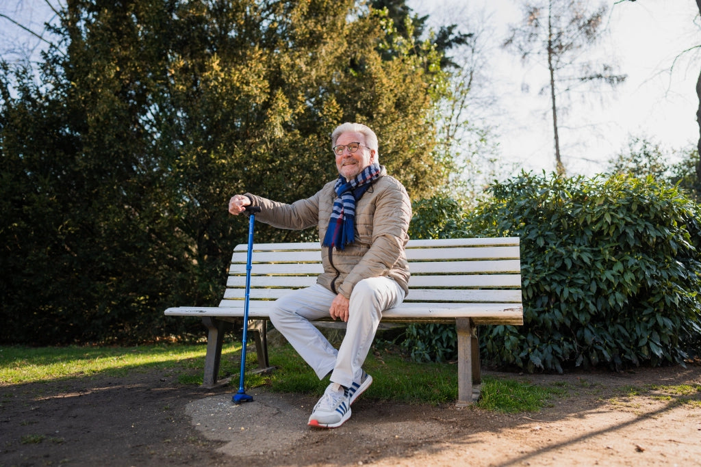 Älterer Mann sitzt auf einer Parkbank und stützt sich auf einen blauen Hurrycane Gehstock von Drive Medical – ideal für sicheres Gehen im Freien.