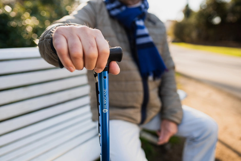 Älterer Mann sitzt auf einer Parkbank und stützt sich auf einen blauen Hurrycane-Gehstock mit ergonomischem Griff.