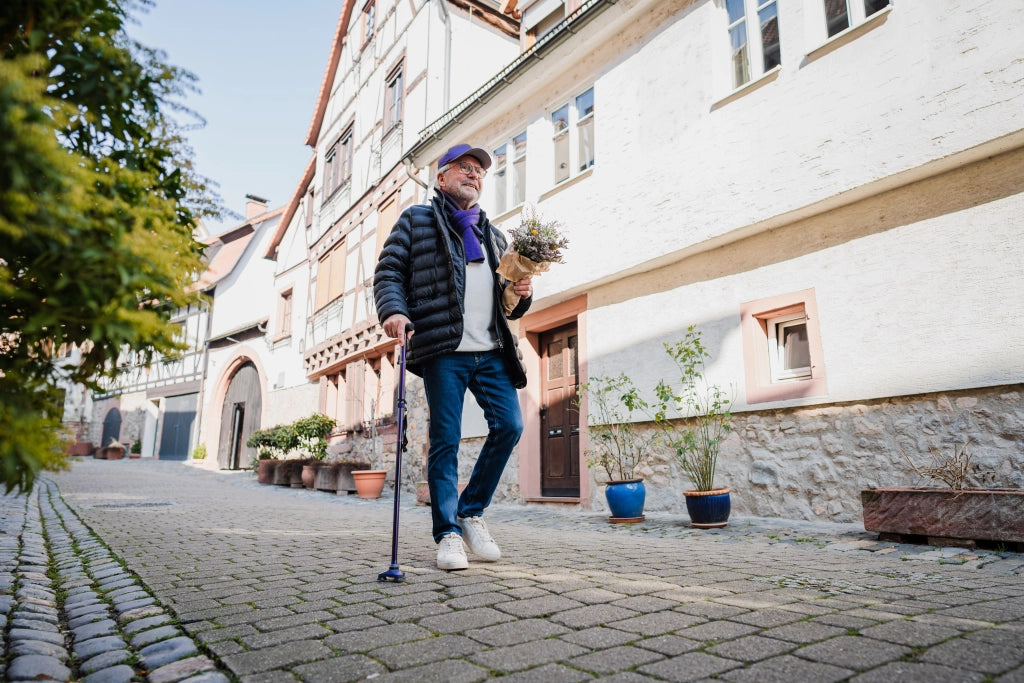 Älterer Mann spaziert mit einem lilafarbenen Gehstock Hurrycane von Drive Medical durch eine Altstadtgasse und trägt einen Blumenstrauß – Mobilität und Stil vereint.