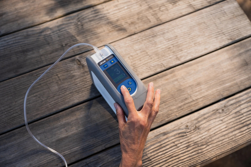 Hand aktiviert den tragbaren Sauerstoffkonzentrator iGo2 von Drive Medical auf einem Holztisch im Freien – perfekte mobile Lösung für die Sauerstofftherapie unterwegs.