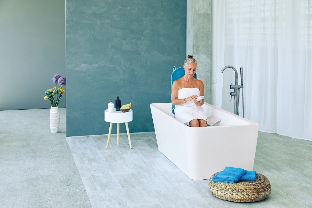 Eine ältere Frau sitzt entspannt in einer freistehenden Badewanne auf einem Badewannenlift mit blauer Rückenlehne und nutzt eine Fernbedienung. Das stilvolle Badezimmer ist modern eingerichtet.