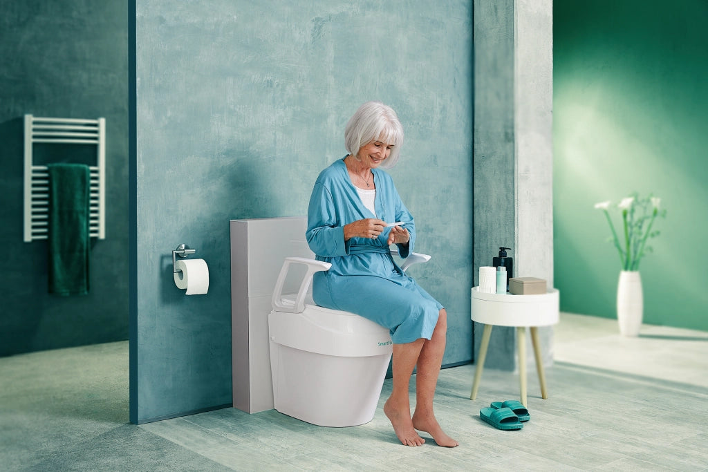 Ältere Frau sitzt entspannt auf der Toilettensitzerhöhung SmartFix B-Protect von Dietz in einem modernen Badezimmer – komfortable Nutzung mit hochklappbaren Armlehnen.
