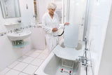 Ältere Frau im Bademantel steht in einem modernen, barrierefreien Badezimmer und bedient mit einer Handsteuerung einen Badewannenlift. Der Lift ist in der Badewanne positioniert, mit einer ergonomischen Rückenlehne und rutschsicherer Sitzfläche, beide mit einem waschbaren Komfortbezug ausgestattet. Die ausklappbaren Seitenflügel erleichtern das Einsteigen. Ideal für Senioren oder Personen mit eingeschränkter Mobilität für ein sicheres und komfortables Badeerlebnis.