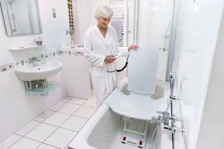 Ältere Frau im Bademantel steht in einem modernen, barrierefreien Badezimmer und bedient mit einer Handsteuerung einen Badewannenlift. Der Lift ist in der Badewanne positioniert, mit einer ergonomischen Rückenlehne und rutschsicherer Sitzfläche, beide mit einem waschbaren Komfortbezug ausgestattet. Die ausklappbaren Seitenflügel erleichtern das Einsteigen. Ideal für Senioren oder Personen mit eingeschränkter Mobilität für ein sicheres und komfortables Badeerlebnis.