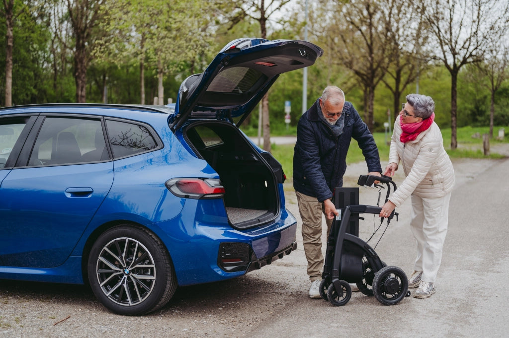 lteres Paar verstaut den zusammengeklappten Rollz Motion Electric im Kofferraum eines blauen Autos. Die Szene zeigt die einfache Transportfähigkeit des faltbaren 3-in-1-Rollators und Elektrorollstuhls, ideal für unterwegs.