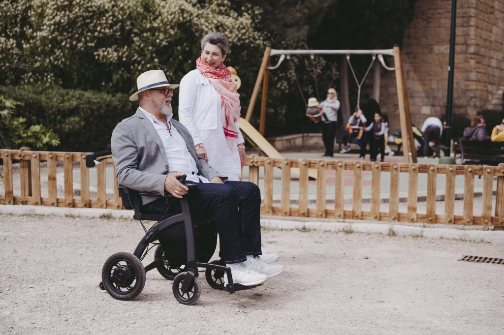 Älterer Mann mit Hut und Sonnenbrille sitzt entspannt im Rollz Motion Electric Elektrorollstuhl, während eine Frau neben ihm geht. Die Szene spielt sich in einem Park mit Spielplatz im Hintergrund ab, was die Alltagstauglichkeit und Flexibilität des Rollstuhls betont.