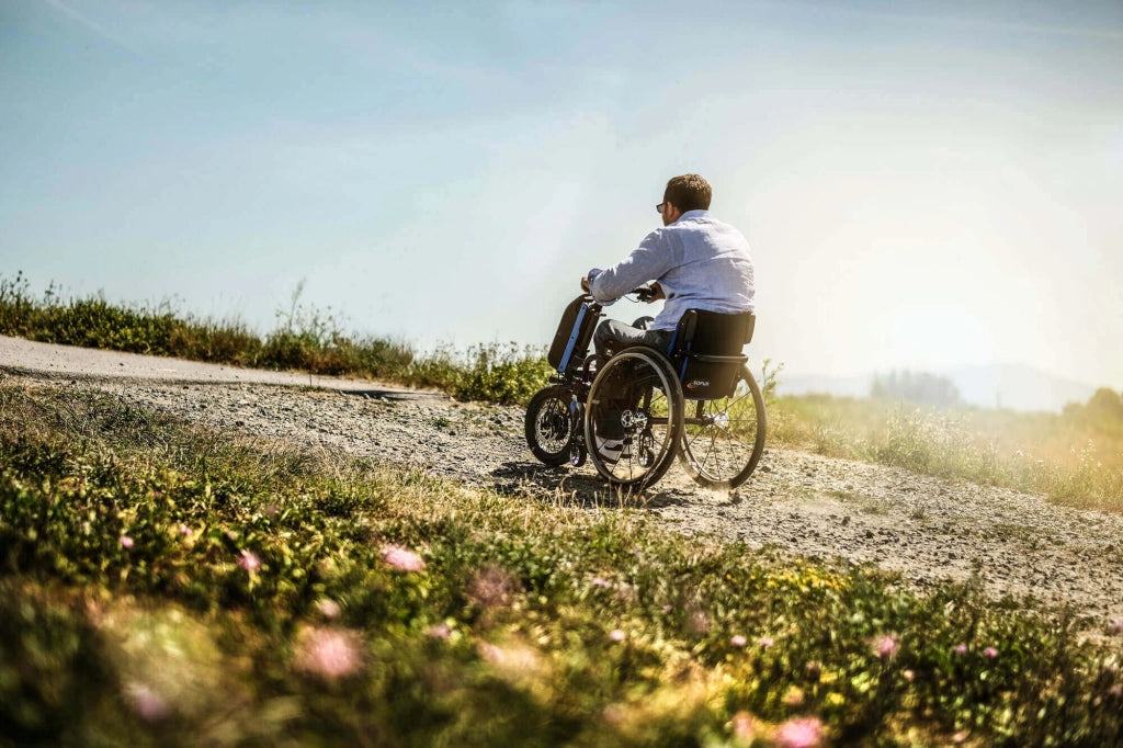 Mann im Rollstuhl mit angedocktem Empulse F55 Rollstuhlzuggerät fährt auf einem Schotterweg durch eine sonnige Landschaft – Symbol für Freiheit und Mobilität im Gelände.