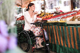 Frau im Rollstuhl mit angedocktem Empulse F55 Rollstuhlzuggerät beim Einkaufen auf dem Wochenmarkt, umgeben von frischem Obst und Gemüse.